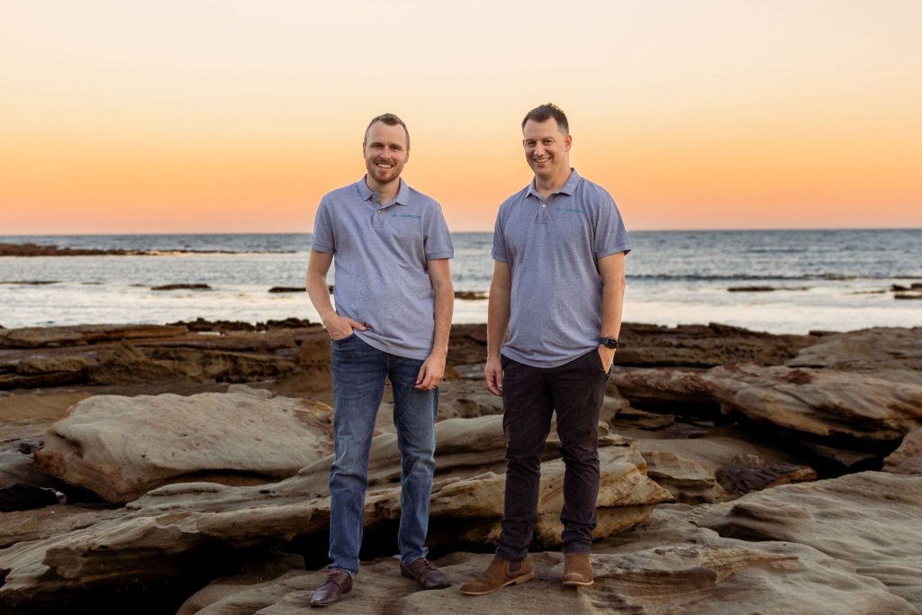 Wade and Justin, directors of Shoreline Lending, standing on coastal rocks at sunset on the Central Coast